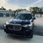 Sleek black SUV professionally detailed by AYB Detailing, featuring a prominent grille and shining headlights, parked on a concrete surface with industrial-style buildings in the background under a clear dusk sky, showcasing exterior auto detailing and gloss enhancement.
