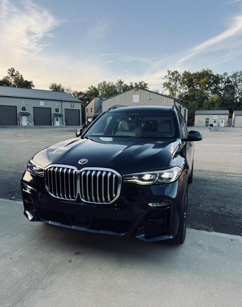 Sleek black SUV professionally detailed by AYB Detailing, featuring a prominent grille and shining headlights, parked on a concrete surface with industrial-style buildings in the background under a clear dusk sky, showcasing exterior auto detailing and gloss enhancement.