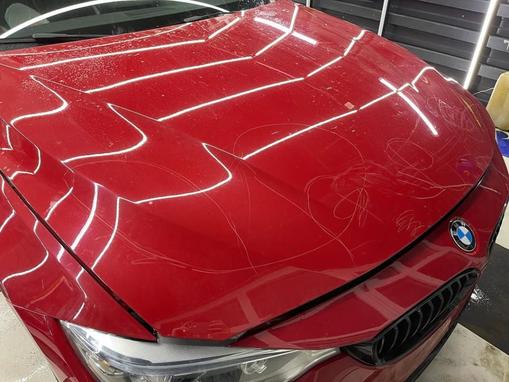 Close-up of a red BMW hood showing scratches before professional enhanced gloss finish and detailing by AYB Detailing.