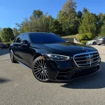 Sleek black Mercedes-Benz luxury car professionally detailed by AYB Detailing, featuring tinted windows and a shiny chrome grille, parked on a sunny day with trees and other vehicles in the background, showcasing exterior detailing and headlight restoration services.
