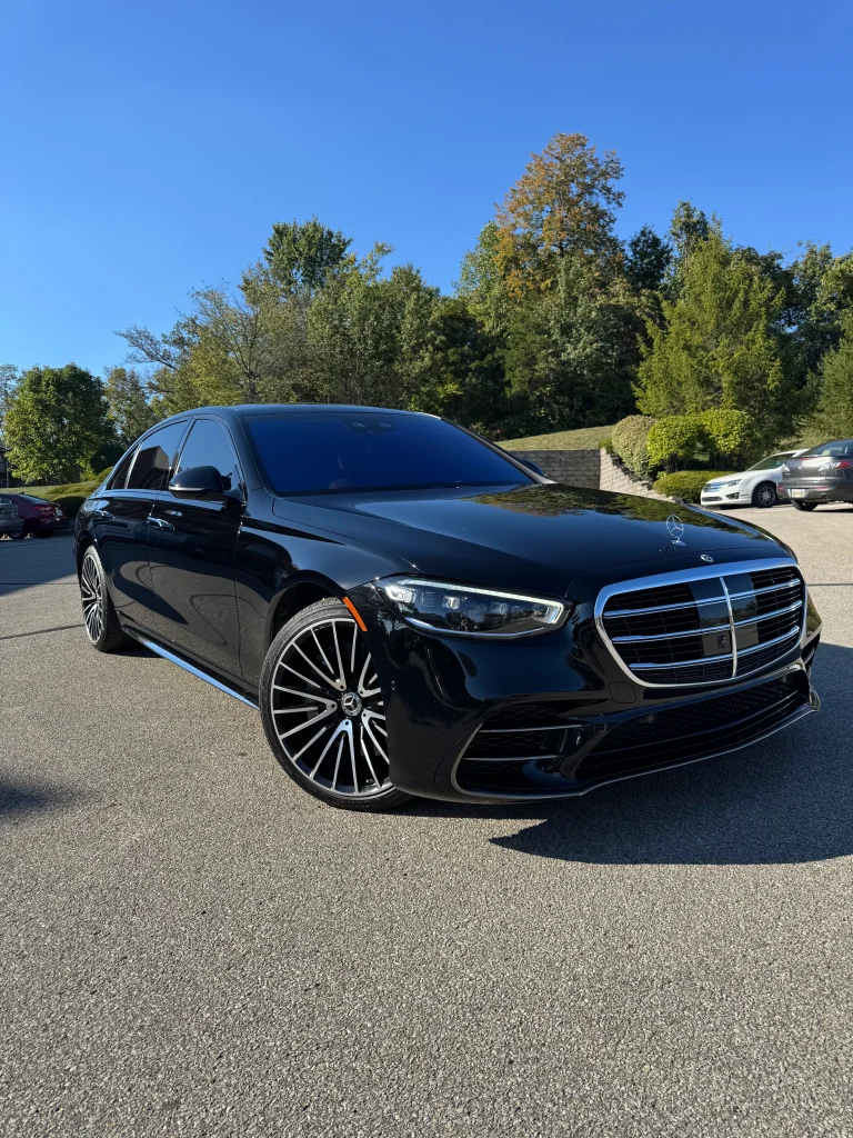 Sleek black Mercedes-Benz luxury car professionally detailed by AYB Detailing, featuring tinted windows and a shiny chrome grille, parked on a sunny day with trees and other vehicles in the background, showcasing exterior detailing and headlight restoration services.