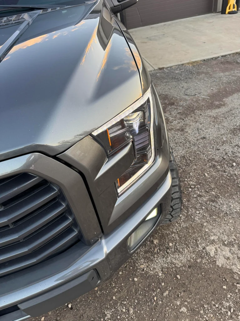 Close-up of a gray pickup truck front professionally detailed by AYB Detailing, highlighting a modern headlight design and chrome grille with a clean, reflective exterior finish under a clear sky.